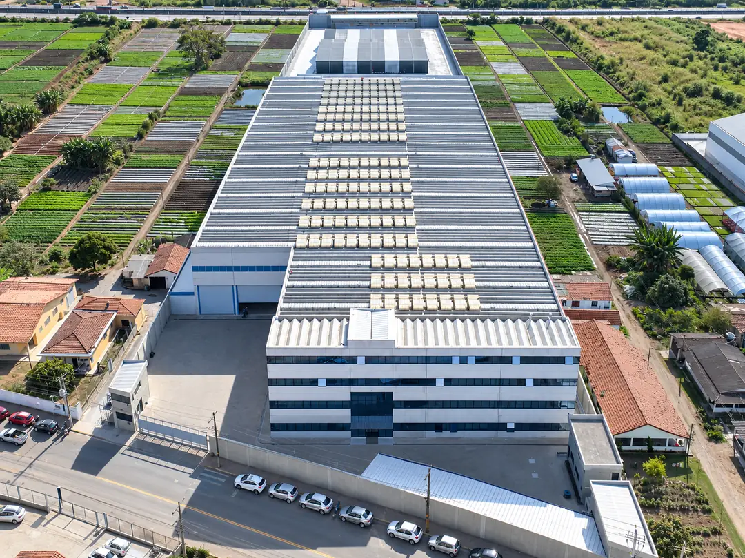Vista aérea de drone do centro de distribuição: galpão comprido de telhado claro, prédio administrativo envidraçado na frente e caminhões no pátio